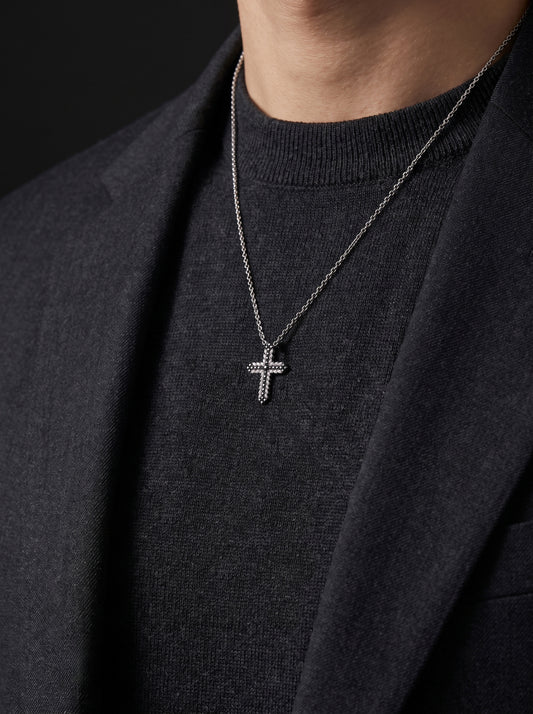 A white gold cross pendant necklace with small clear stones, worn on a model's neck over a dark gray knit sweater and dark blazer.