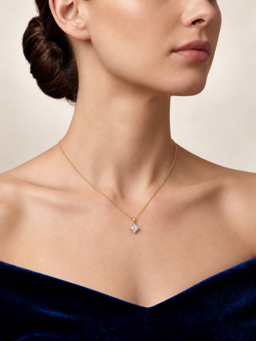 A close-up of a model wearing a delicate gold chain necklace with a small diamond pendant. The model is wearing a dark blue velvet off-the-shoulder top against a neutral beige background.