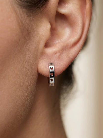 Close-up of a platinum huggie earring worn on the ear, showing heart-shaped cut-outs across polished facets.