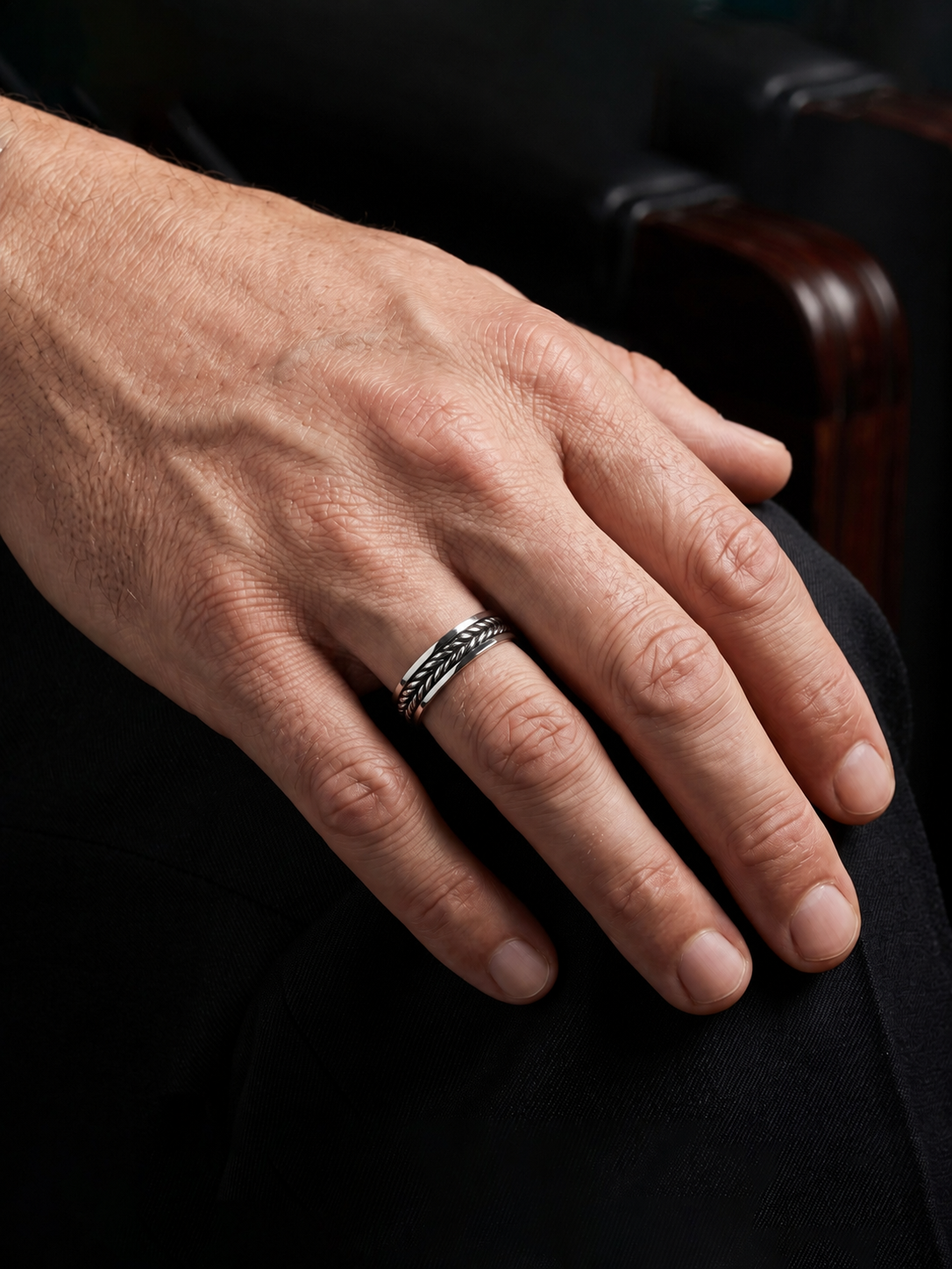 A close-up of a man's hand resting on his knee, wearing a platinum ring with a textured, braided band design.