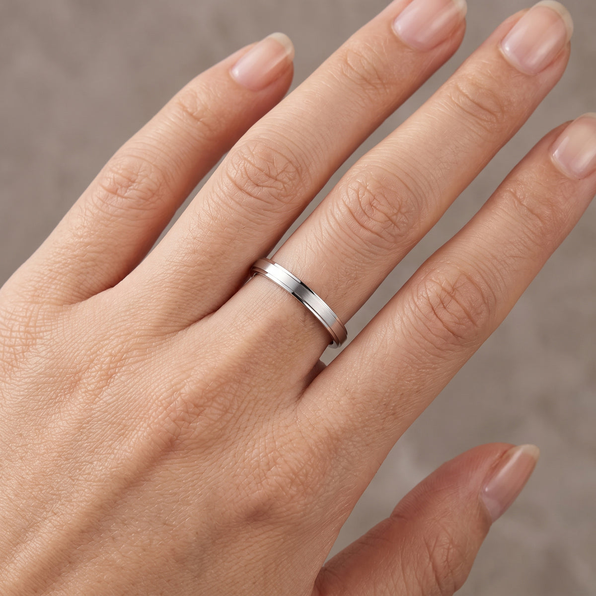 A close-up of a hand wearing a simple polished platinum band ring on one of the middle fingers, set against a soft neutral gray background.